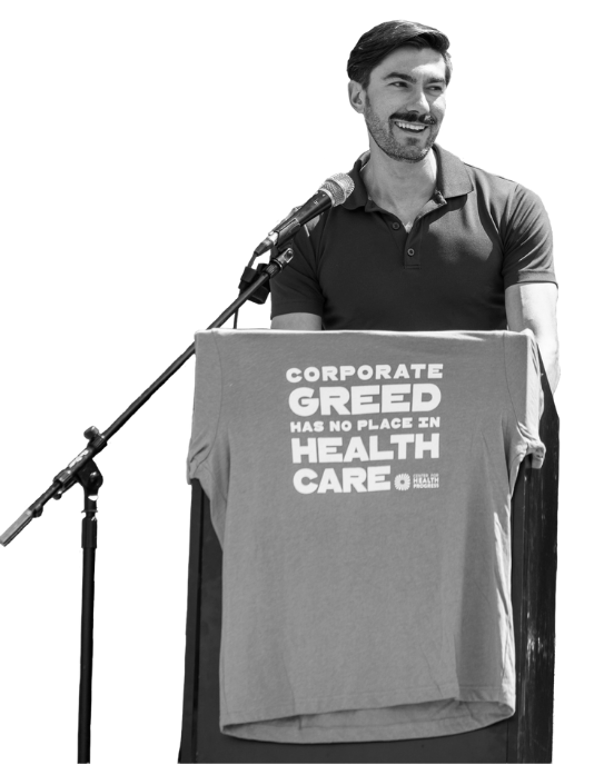 A black and white image of a young light skin man standing and speaking at a podium with a microphone, with a t-shirt draped over the podium that reads “Corporate greed has no place in healthcare.”