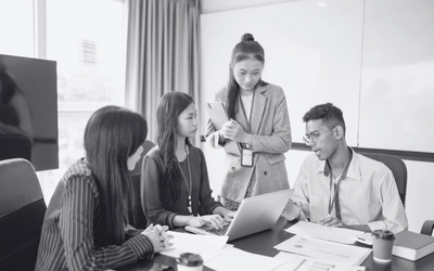A black and white image of a group of four young, professional adults, three of whom are sitting around a table with papers, coffee cups, and looking at a laptop, while one stands above them looking down at the laptop.
