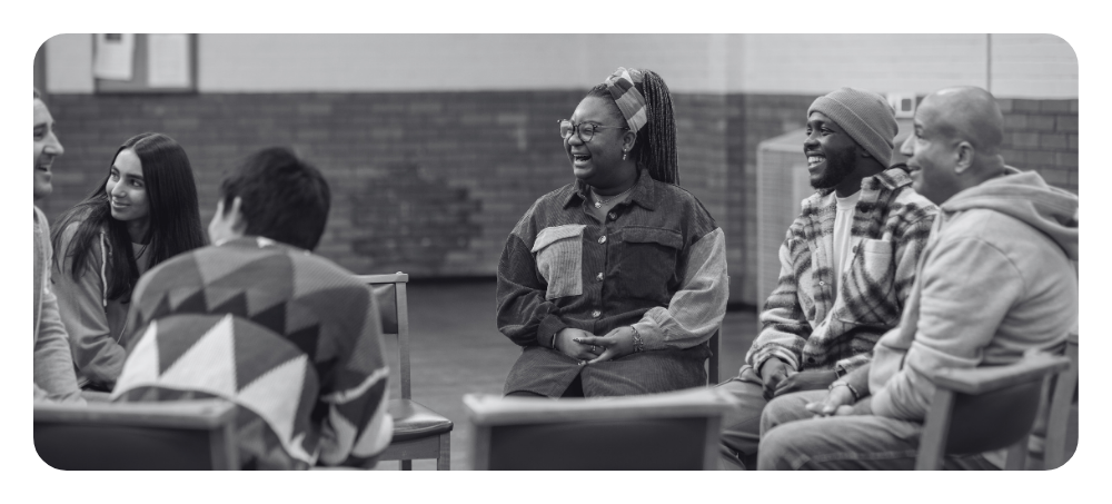 A black and white image of a group of multi-racial and multi-generational people sitting in a circle of chairs, talking and smiling with each other.